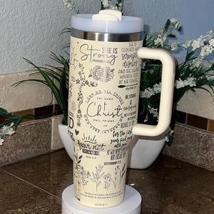 May include: A white and cream stainless steel tumbler with a black handle. The tumbler has a floral design and inspirational quotes with bible verses. The tumbler is on a white rotating base.