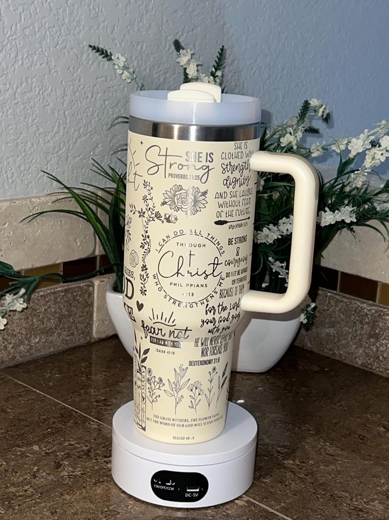 May include: A white and cream stainless steel tumbler with a black handle. The tumbler has a floral design and inspirational quotes with bible verses. The tumbler is on a white rotating base.