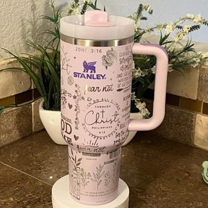 May include: A pink stainless steel travel mug with a black handle. The mug has a variety of inspirational quotes and bible verses printed on it, including "fear not" and "do all things through Christ". The mug is sitting on a white rotating display stand.