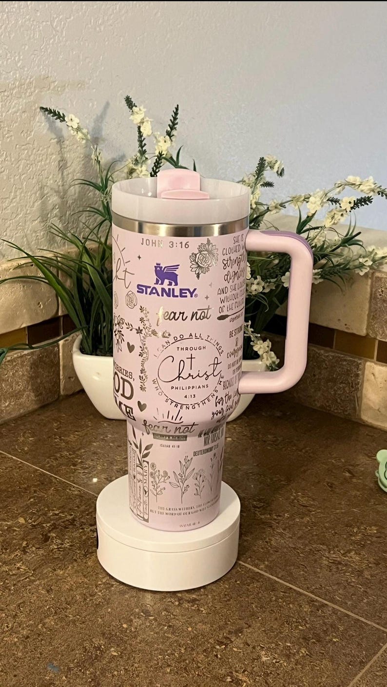 May include: A pink stainless steel travel mug with a black handle. The mug has a variety of inspirational quotes and bible verses printed on it, including "fear not" and "do all things through Christ". The mug is sitting on a white rotating display stand.