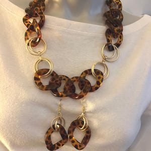 May include: A chunky necklace and matching drop earrings set featuring brown and black leopard print resin links interspersed with textured gold-tone metal rings. The necklace consists of large oval links, while the earrings feature a single matching resin link suspended from a gold-tone ring.