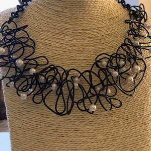 May include: A black wire necklace with white pearls. The necklace is in a loose, organic shape, and the pearls are scattered throughout the wire.