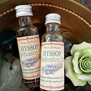 May include: Two small glass bottles with silver lids filled with a clear liquid. The bottles have labels that say "Silvana Apothecary Hyssop Purification Oil" and include a quote about hyssop. The bottles are sitting on a gold surface with a green rose.