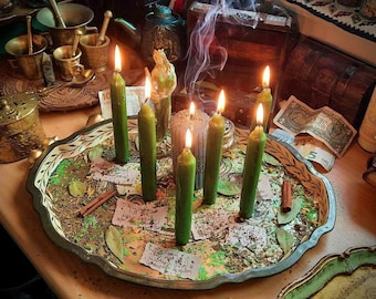 May include: A green, leafy, circular tray with six green candles, a silver candle holder, and cinnamon sticks. The tray is on a wooden table with other items, including a small gold box, a book, and a few bills.