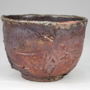 May include: A handcrafted ceramic bowl with a rustic, textured finish. The bowl is a dark reddish-brown colour with lighter brown and grey accents. The rim has a slightly uneven, wavy edge, and the exterior has carved patterns.