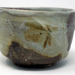 May include: A ceramic teacup with a light brown, grey, and purple glaze. The cup has an uneven, textured surface and a slightly rounded shape.