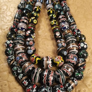 May include: Strands of colorful glass beads in various patterns. The beads are primarily black with accents of red, white, yellow, and green. Some beads have striped or floral designs, while others have metallic accents. The beads are strung together, creating a necklace.
