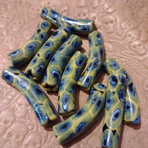 May include: A collection of handmade, curved beads. Each bead features a yellow base with blue and black circular patterns. The beads are arranged on a textured, light brown surface, showcasing their unique design and color variations.