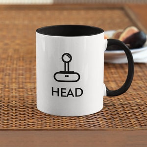 May include: White ceramic mug with a black rim and handle. The mug features a black and white illustration of a joystick with the word "HEAD" printed below it.