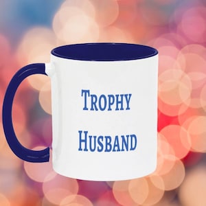 Peut inclure: Mug en céramique blanche avec intérieur et anse bleu marine. Le mug porte l'inscription "Trophy Husband" en bleu, centrée sur le devant. L'arrière-plan est un mélange flou de rose, de rouge et de bleu.