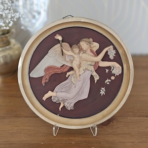 May include: A circular wall plaque featuring a raised relief of a winged figure carrying a small child against a dark maroon background. The figures are rendered in cream and soft pink tones, with the winged figure holding a torch and flowers.