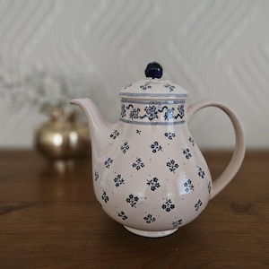 May include: A ceramic teapot with a lid, featuring a light pink base color and a pattern of small blue floral designs. The lid and upper rim have a blue and white decorative border. The teapot has a curved handle and a spout.