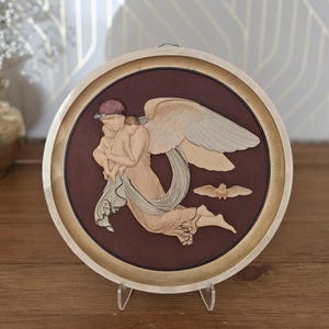 May include: A circular wall plaque featuring a relief sculpture of a winged figure holding a small child against a dark maroon background. The figure has light-colored skin and flowing drapery, with a small bird flying nearby, all encased in a light wood frame.