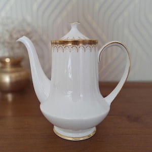 May include: A white porcelain coffee pot featuring a tall, slender body, a curved handle, and a long spout. The rim of the lid and the upper section of the pot are decorated with an intricate gold filigree pattern. A thin gold band accents the base.