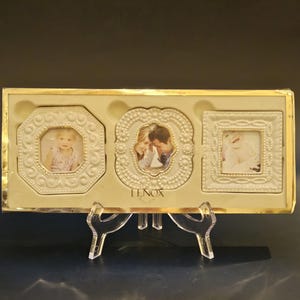 May include: A Lenox triple photo frame set with three ivory-coloured frames in a gold-toned box. The frames have ornate detailing and hold small photos. The set is displayed on a clear acrylic stand.