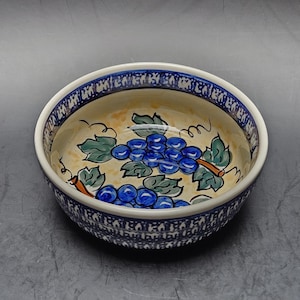 May include: A round ceramic bowl featuring a hand-painted design of blue grapes with green leaves on a cream-colored background. The rim and exterior are decorated with a repeating blue and white geometric pattern on a dark blue border.