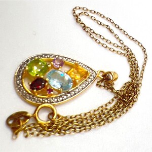 May include: A gold-toned pendant necklace featuring a teardrop-shaped pendant. The pendant is adorned with an array of colorful gemstones, including green, blue, red, and yellow stones, set within a gold frame. The pendant is accented with small, clear stones around the edge.