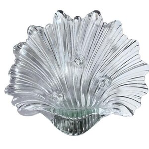 May include: Clear glass seashell-shaped decorative bowl. The bowl has a scalloped edge and three small, round, decorative elements. The bowl is designed with vertical lines that create a fan-like pattern.
