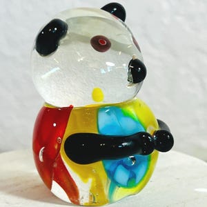 May include: A colorful glass panda figurine with a clear head, black ears and eyes, and a body in red, yellow, and blue. The panda's arms are black, and it has a small yellow spot on its chest. This decorative item is a unique piece of art.
