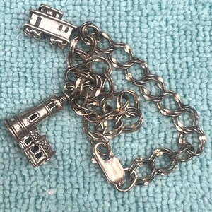 May include: A silver-toned charm bracelet featuring a cable chain with a lobster claw clasp. The bracelet includes two dangling charms: one shaped like a classic trolley car and another detailed charm depicting a lighthouse structure.