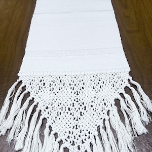 May include: White table runner with a woven design and fringe. The runner has a simple, elegant design and would be perfect for a casual or formal dining setting.