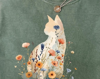 May include: A moss green t-shirt with a cat silhouette filled with colorful flowers. The shirt has a Comfort Colors tag and a silver necklace with a small pendant. The word "Moss" is in the upper left corner.