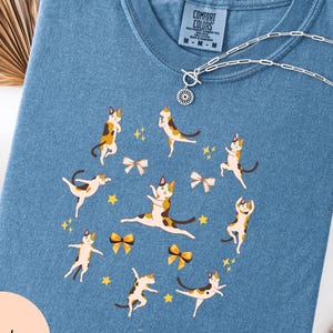 May include: Blue jean coloured t-shirt with a crew neck and short sleeves. The shirt features a playful design of calico cats in various poses, surrounded by yellow bows and stars. The shirt has the text "Comfort Colors" on the tag.