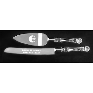 May include: A set of two silver cake knives with clear crystal handles. The top knife has a Rebel Alliance symbol engraved on the blade. The bottom knife has the text "Linda & Dennis March 1, 2023" engraved on the blade.