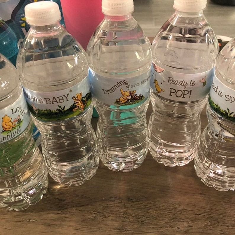 May include: Four clear plastic water bottles with Winnie the Pooh themed labels. The labels have blue backgrounds and feature Winnie the Pooh and Piglet. The labels read "Love is in the air", "Welcome Baby", "Dreaming of a new little one", and "Ready to POP!"
