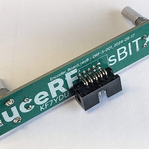 Puede incluir: Una placa de circuito verde con el texto "duceRF" y "sBITX". Tiene dos perillas plateadas en cada extremo y un conector negro en el medio. La placa también tiene el texto "Encoder Board_revB - DRF-S-001_2024-05-17".