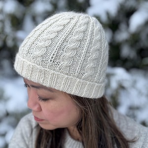 Puede incluir: Un gorro de punto color crema con un diseño de punto trenzado. El gorro tiene un borde acanalado y una parte superior redondeada. El gorro está hecho de un hilo suave y texturizado, adecuado para el clima frío.