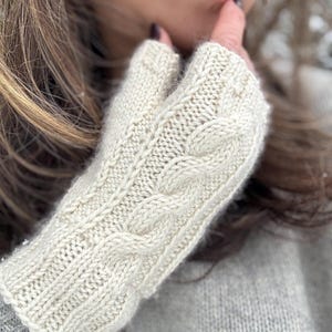 Puede incluir: Guantes sin dedos de punto color crema con diseño de punto trenzado. Los guantes están hechos de un hilo suave y texturizado y están diseñados para mantener las manos calientes y permitir el movimiento de los dedos. Los guantes son de color neutro.
