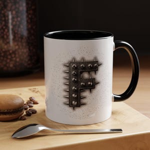 May include: White ceramic mug with a black interior and handle. The mug features a studded, metallic letter "F" design. The mug is on a wooden surface with a spoon and a pastry.