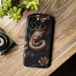 May include: A black phone case with a gold snake and floral design. The snake has a red eye and is coiled around a branch. The background is dark and the flowers are gold.