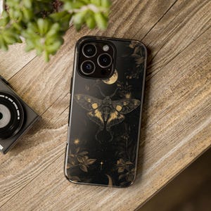 Puede incluir: Una funda de teléfono negra con un diseño de polilla dorada y floral sobre un fondo oscuro. La funda tiene un patrón de luna y estrellas y un recorte para el objetivo de la cámara.