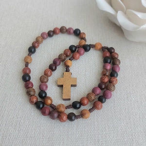 May include: A rosary featuring wooden beads in various shades of brown, black, and purple. The beads are connected by a brown cord, with a wooden cross pendant. The rosary is presented on a neutral-toned background.