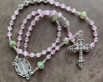 May include: A pink and green rosary with a silver cross and a silver charm featuring the Virgin Mary. The rosary beads are made of glass and have a frosted finish. The charm has a floral design and the words "KAYLA" are spelled out on the beads.
