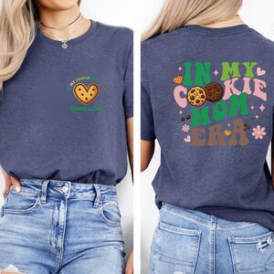 a woman wearing a blue t-shirt with a graphic design on the back. The design includes a heart and cookies, along with the text "In my cookie mom era".