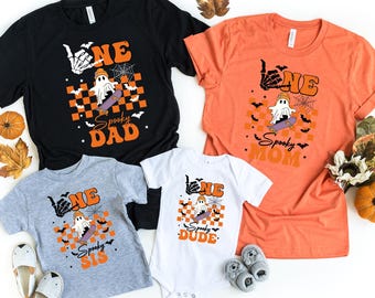 May include: Four Halloween-themed t-shirts and a onesie in black, orange, gray, and white. Each shirt features a ghost on a skateboard, the word "ONE", and the phrases "Spooky Dad", "Spooky Mom", "Spooky Sis", and "Spooky Dude".
