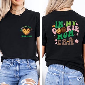 a woman wearing a black t-shirt with a graphic design on the back. The design includes a heart-shaped cookie and the text "In my cookie mom era" along with other cookie-related imagery.
