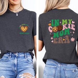 a woman wearing a gray t-shirt with a graphic design on the back. The design includes a cookie and the text "In my cookie mom era" in a colorful, playful style.