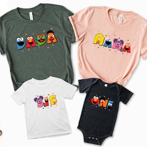 a collection of four t-shirts, each featuring a different character from the Sesame Street television series. The characters are arranged in a way that suggests they are part of a family, with the first shirt on the left, the second shirt on the right, and the third shirt in the center. The fourth shirt is a baby onesie, which is placed on the bottom right corner of the image.