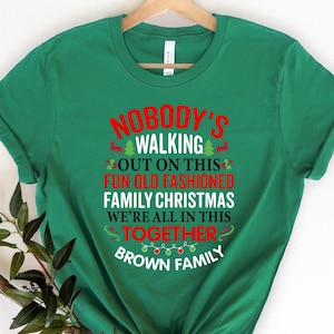 May include: Green t-shirt featuring the text "NOBODY'S WALKING OUT ON THIS FUN OLD FASHIONED FAMILY CHRISTMAS WE'RE ALL IN THIS TOGETHER BROWN FAMILY" in red and white. The shirt has a crew neck and short sleeves.
