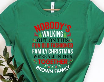 May include: Green t-shirt with the text "NOBODY'S WALKING OUT ON THIS FUN OLD FASHIONED FAMILY CHRISTMAS WE'RE ALL IN THIS TOGETHER BROWN FAMILY" in red and white. The shirt has a crew neck and short sleeves.