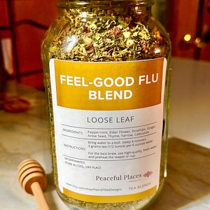May include: A glass jar filled with loose leaf tea, labeled "FEEL-GOOD FLU BLEND." The label lists ingredients like peppermint and elderflower. A wooden honey dipper rests beside the jar. The tea is decaffeinated and the jar is sealed with a gold lid.
