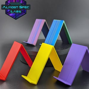 May include: A collection of colorful, angled phone stands in red, yellow, and purple. Two stands feature striped patterns in blue and purple. The stands are arranged on a dark surface, with the text "ALMOST-SPEC LABS" visible in the background.