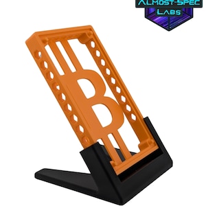 May include: An orange Bitcoin symbol phone stand with a black base. The stand is made of plastic and features the Almost-Spec Labs logo in the upper right corner. The Bitcoin symbol is prominently displayed in the center of the stand.