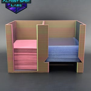 Pode incluir: Um porta-cartões com vários compartimentos com pilhas de cartões rosa e azuis. O suporte é feito de um material iridescente texturizado com o texto "ALMOST-SPEC LABS" no canto superior esquerdo.