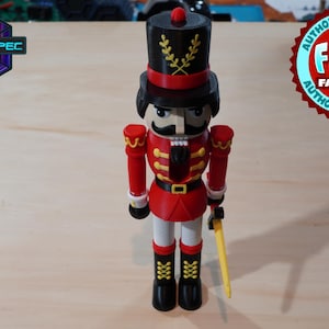 Puede incluir: Un cascanueces de juguete rojo y dorado con un sombrero negro y un bigote. El cascanueces sostiene un palo dorado en su mano derecha.