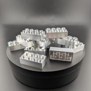 以下が含まれることがあります： 複数の丸いスタッドが上部に、3つの丸い穴が下部にある銀色のブロックのコレクション。ブロックは黒い表面に配置され、背景は暗いです。